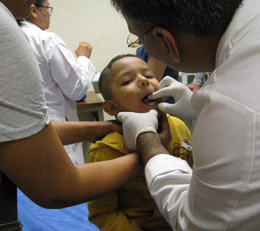 A doctor wearing gloves examines a young boy’s mouth while another adult gently holds him still. Medical staff are visible in the background at SunCoast, a trusted name in plastic surgery in Houston.