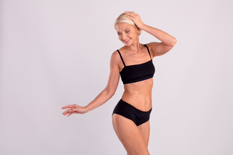 A smiling older woman with short blond hair poses confidently in a black bikini against a plain light background, embracing her beauty after plastic surgery in Houston, with one hand touching her head and the other extended outward.