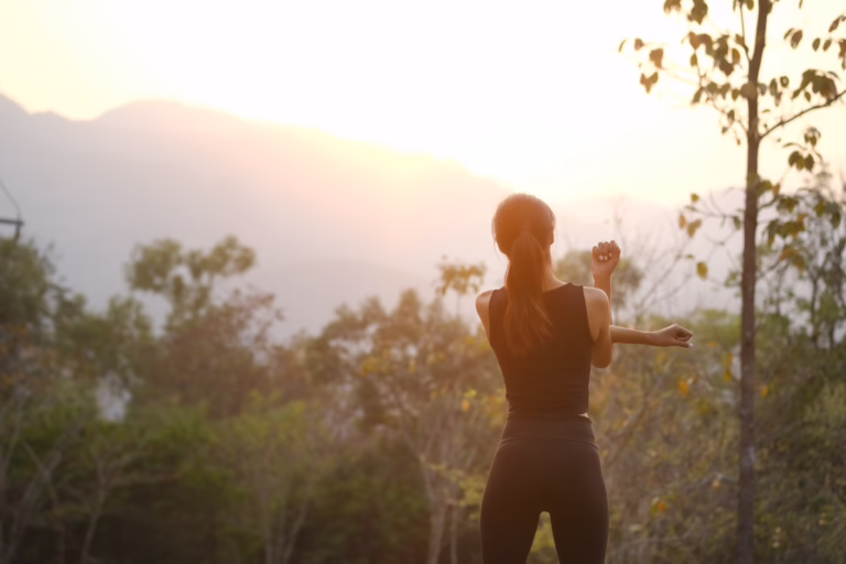 A person with long hair in a ponytail stretches their arm outdoors at sunrise or sunset, surrounded by trees and mountains, basking in the soft, warm light. The scene captures a peaceful moment reminiscent of Houston’s scenic beauty.
